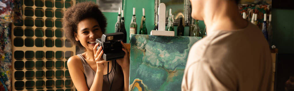 felice donna afroamericana con fotocamera vintage scattare foto di fidanzato sfocato vicino alla pittura su cavalletto, banner - Foto, immagini