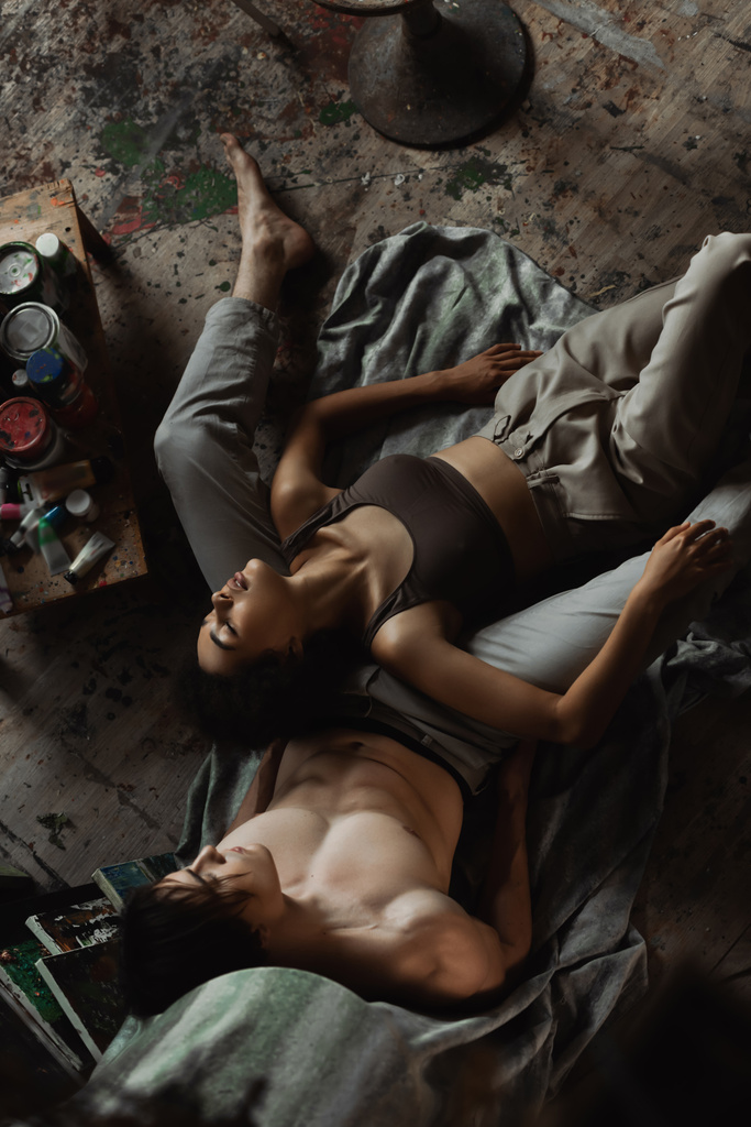 Tutkulu Afrikalı Amerikalı kadın üstte, pantolonu kapalı, gözleri kapalı atölyede seksi bir adamın yanında yatıyor. - Fotoğraf, Görsel