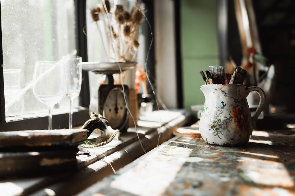handmade jug with paintbrushes near vintage scales and glasses on windowsill in sunlight  - Photo, Image