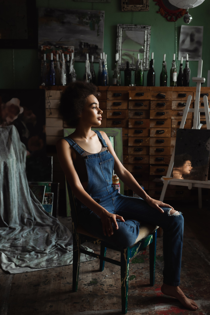 brunette afrikaanse amerikaanse vrouw in overalls zitten in workshop met vintage decor en weg te kijken - Foto, afbeelding