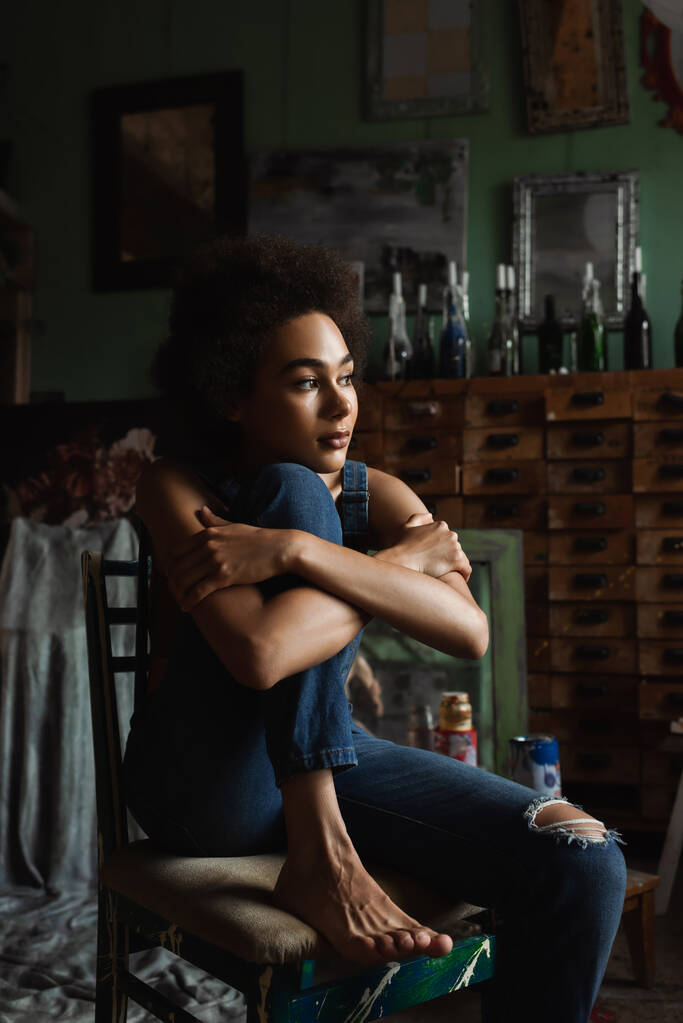 pretty and thoughtful african american woman in overalls sitting with crossed arms in art studio - Photo, Image