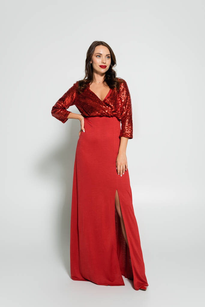Volle Länge der hübschen brünetten Frau im roten Kleid hält Hand auf Hüfte auf grauem Hintergrund - Foto, Bild