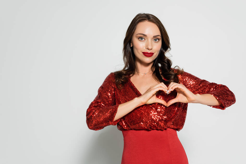 Стильная брюнетка в красном платье показывает жест сердца на сером фоне  - Фото, изображение