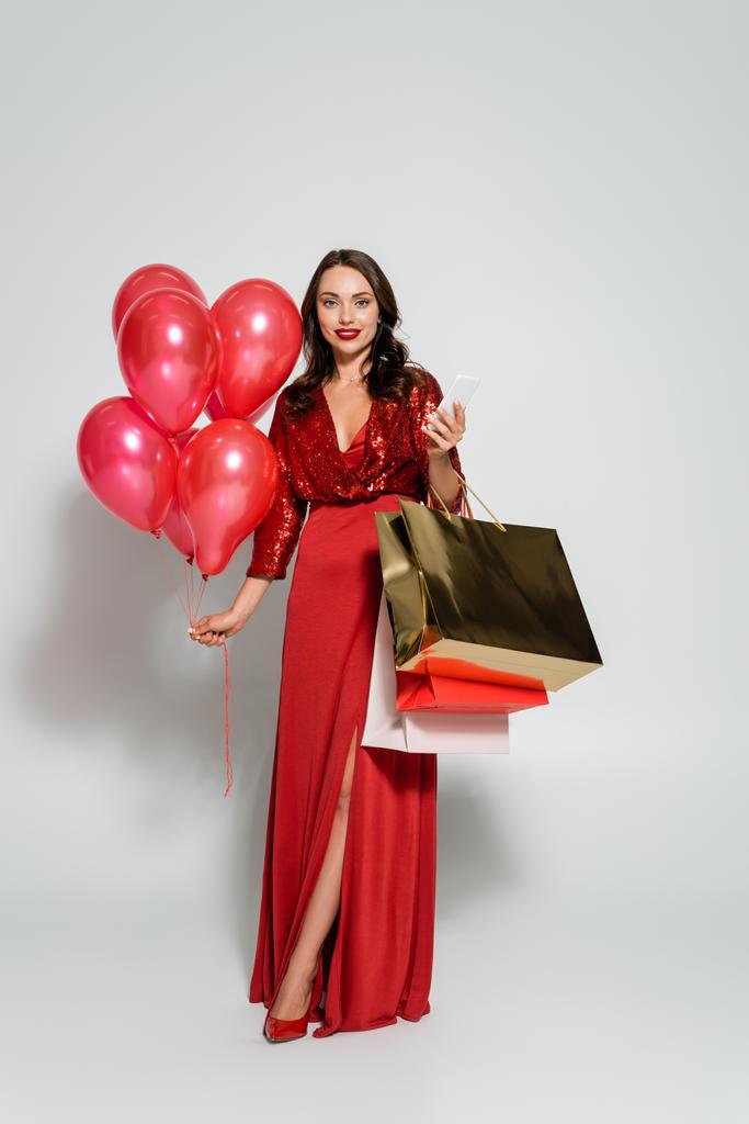 Kırmızı elbiseli, elinde akıllı telefon ve gri arka planda balonlar olan bir kadın.  - Fotoğraf, Görsel