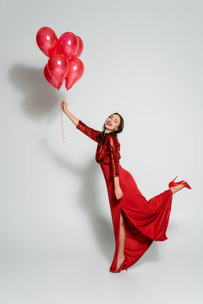 Wesoła młoda kobieta w czerwonej sukience trzymająca balony i patrząca w kamerę na szarym tle  - Zdjęcie, obraz