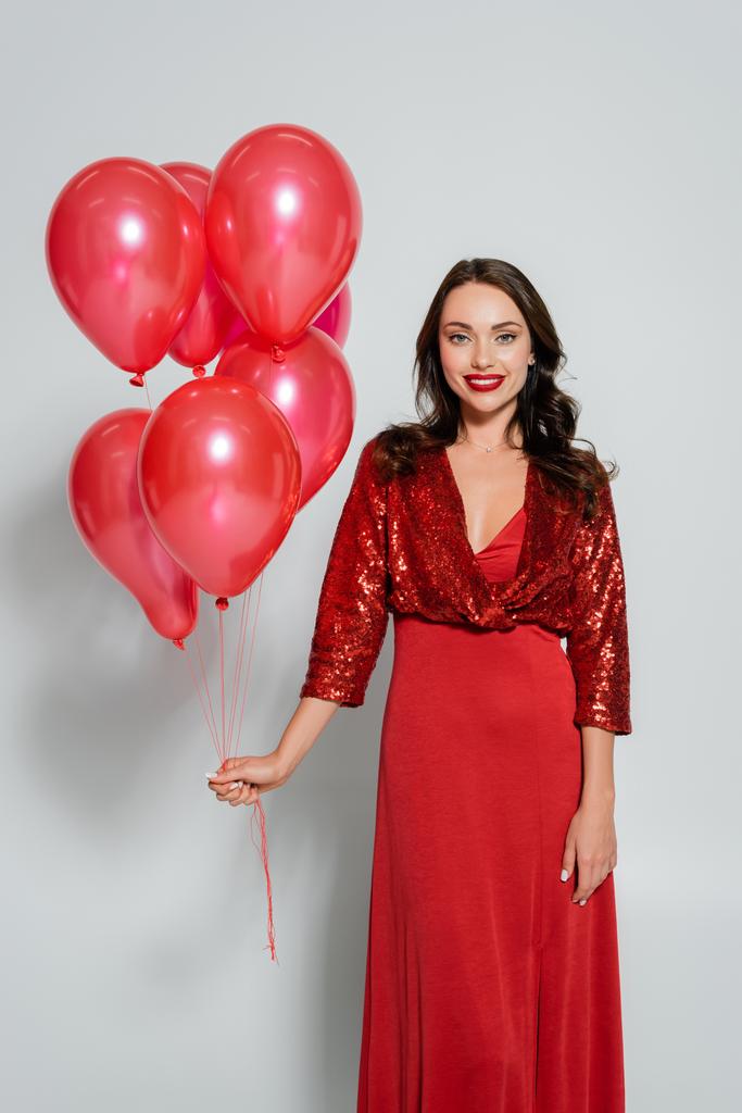 Молодая женщина в красном платье держит праздничные воздушные шары на сером фоне  - Фото, изображение