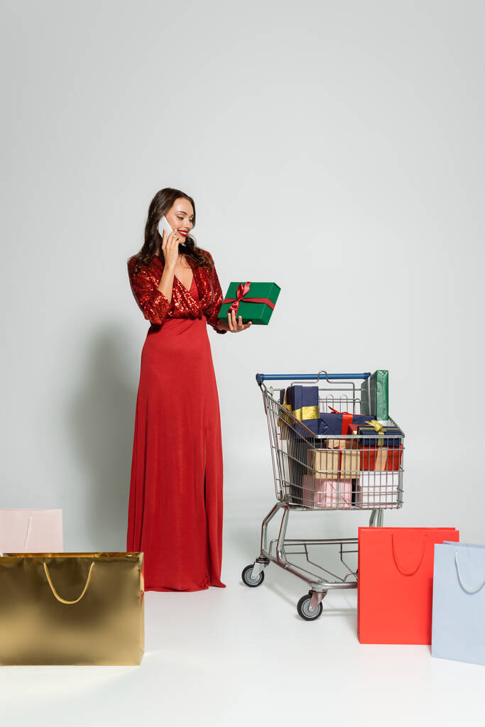 スマートフォンで話すスタイリッシュな女性とグレーの背景にショッピングバッグの近くの贈り物を保持  - 写真・画像