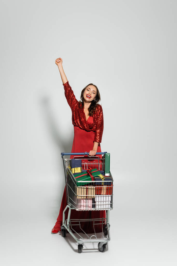 Donna eccitata in abito rosso in piedi vicino carrello della spesa con regali su sfondo grigio  - Foto, immagini
