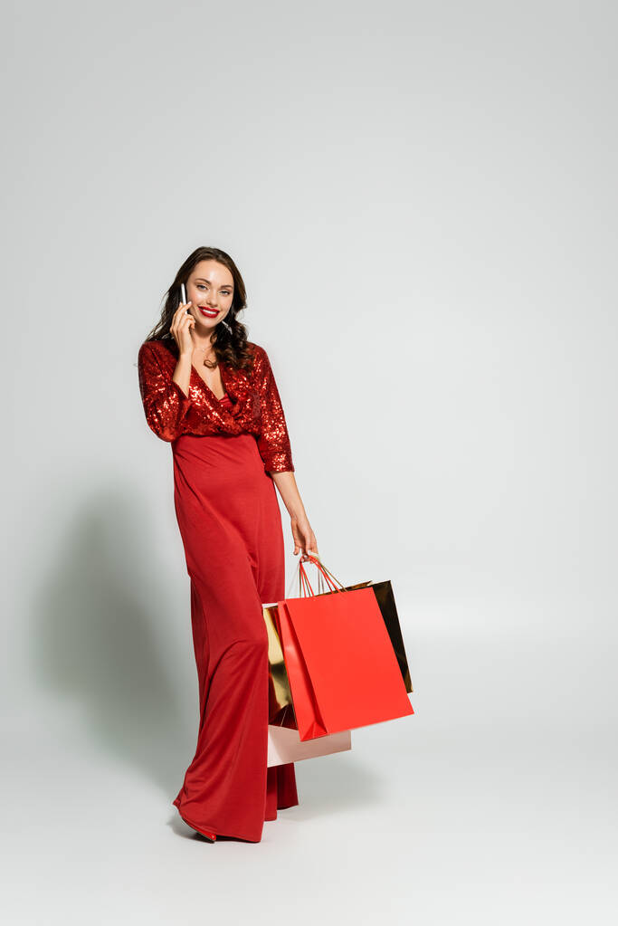 Stílusos nő ruha beszél okostelefon és gazdaság bevásárló táskák szürke háttér  - Fotó, kép