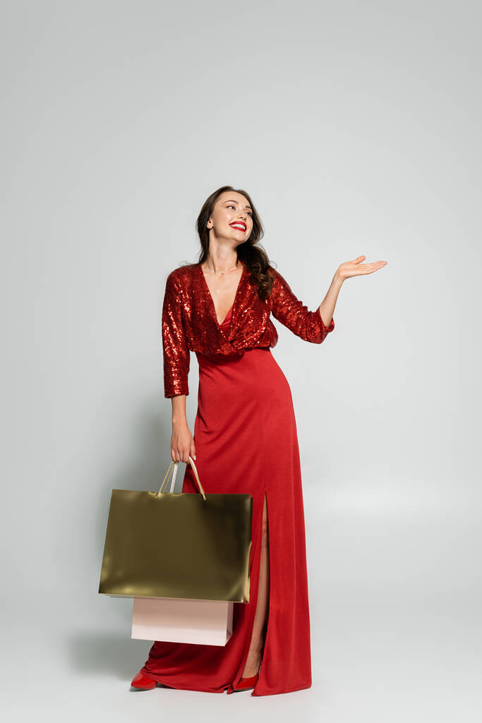 Boldog nő piros ruhában mutató kézzel és gazdaság bevásárló táskák szürke háttér  - Fotó, kép