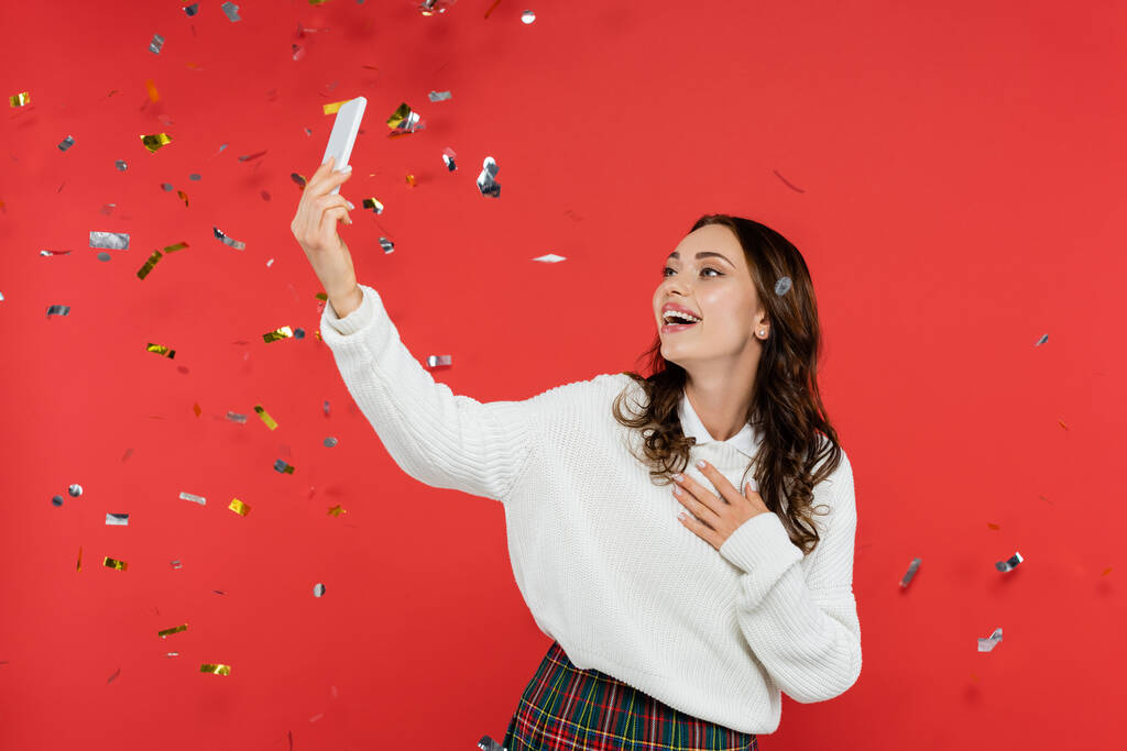 Mujer alegre en suéter tomando selfie bajo confeti sobre fondo rojo  - Foto, imagen