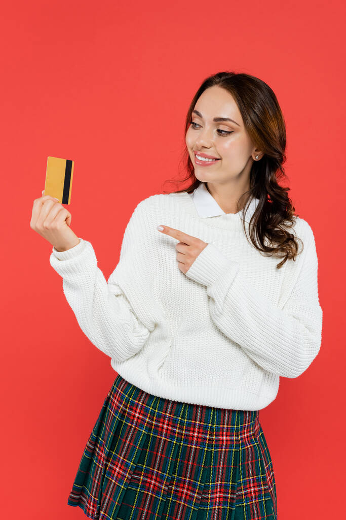 Улыбающаяся женщина в клетчатой юбке и свитере, указывающая на кредитку, изолированную на красном  - Фото, изображение