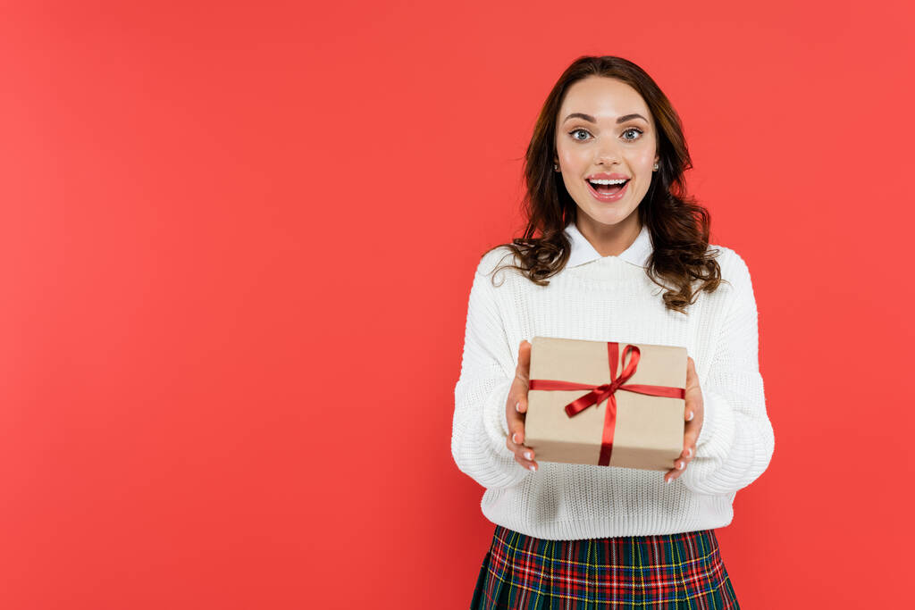 Šťastná mladá žena v jumper držení dárkové krabice a při pohledu na kameru izolované na červené  - Fotografie, Obrázek