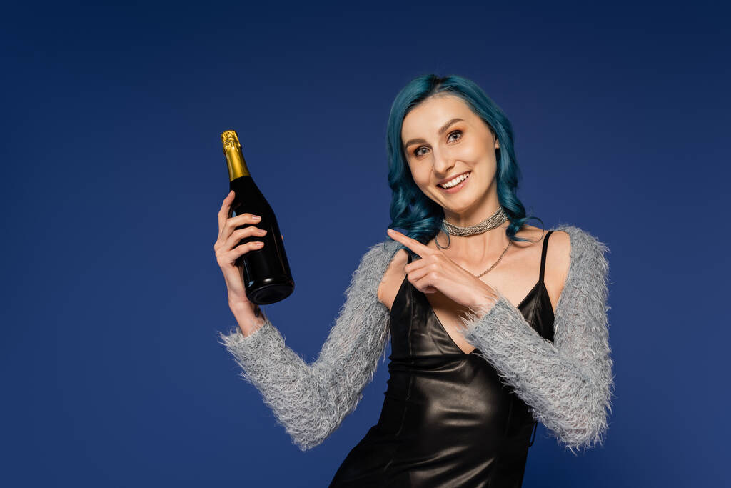 vrolijke vrouw in zwart lederen jurk wijzend naar champagne fles geïsoleerd op blauw - Foto, afbeelding
