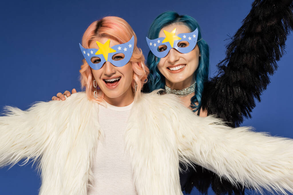 ενθουσιασμένοι queer φίλοι με μάσκες κόμμα και χνουδωτά faux γούνα σακάκια κοιτάζοντας κάμερα που απομονώνονται σε μπλε - Φωτογραφία, εικόνα