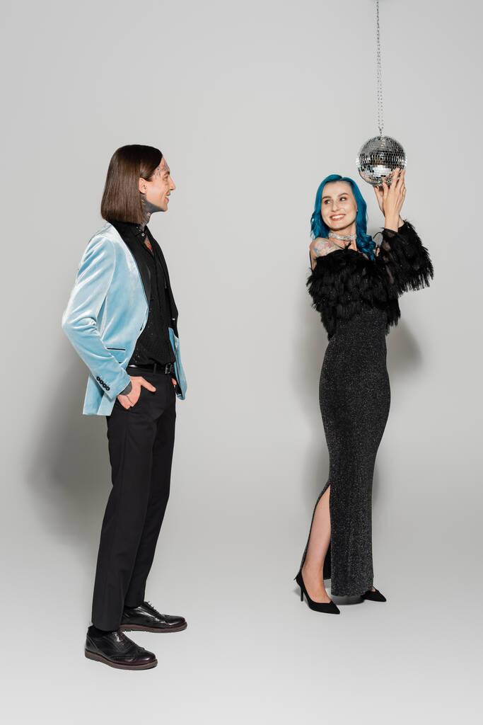 cheerful nonbinary person touching disco ball near queer friend standing with hand in pocket on grey background - Photo, Image