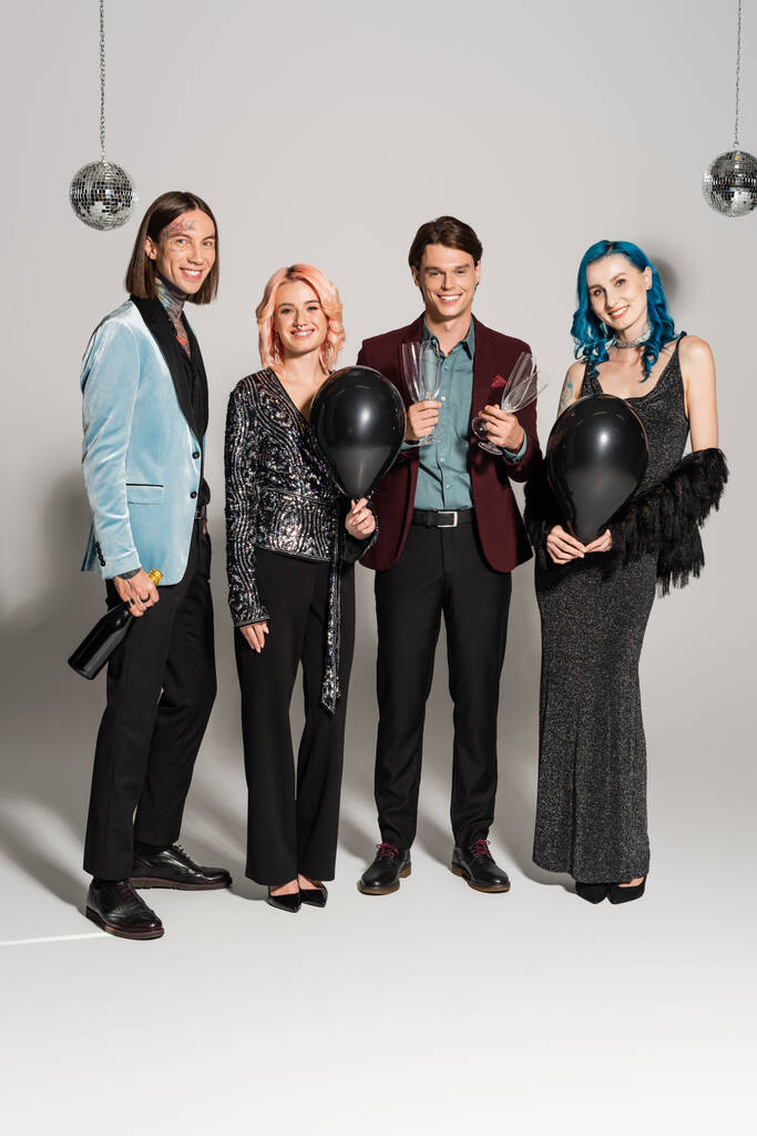 full length of happy queer people in festive clothes holding champagne and black balloons on grey background - Photo, Image