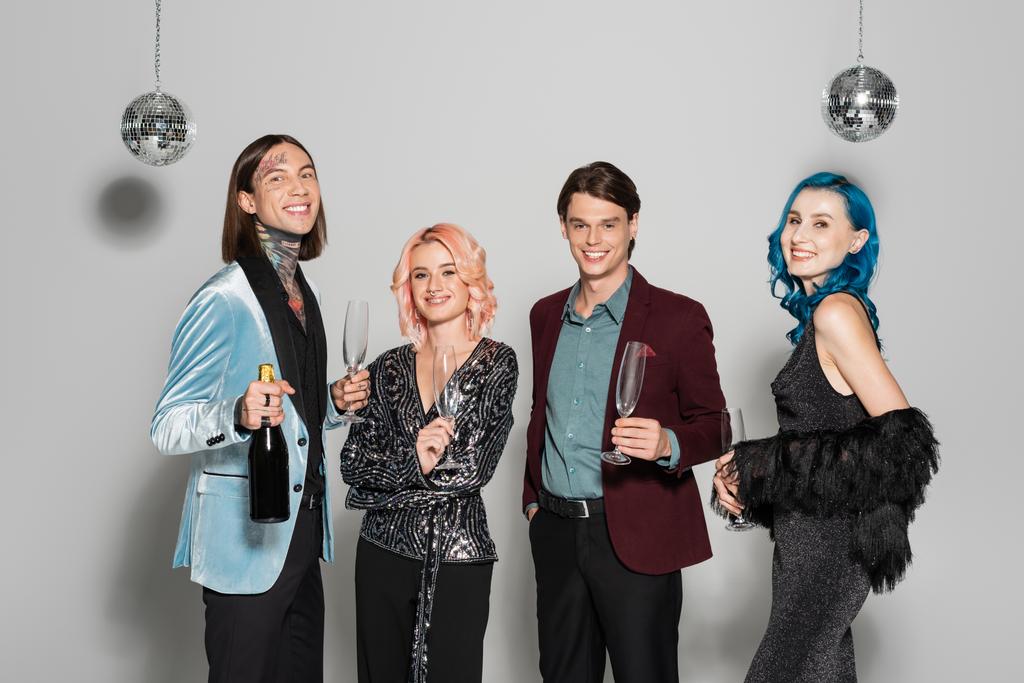 elegante nichtbinäre Freunde mit Champagner lächeln in die Kamera neben Discokugeln auf grauem Hintergrund - Foto, Bild