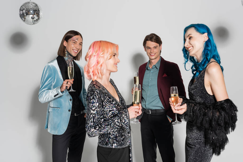 gelukkig niet binaire vrienden met champagne bril glimlachen naar elkaar in de buurt queer mensen op grijze achtergrond - Foto, afbeelding
