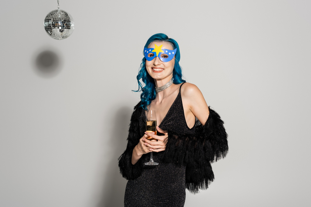 vrouw in elegante kleren en party masker houden champagne en kijken naar de camera op grijze achtergrond - Foto, afbeelding