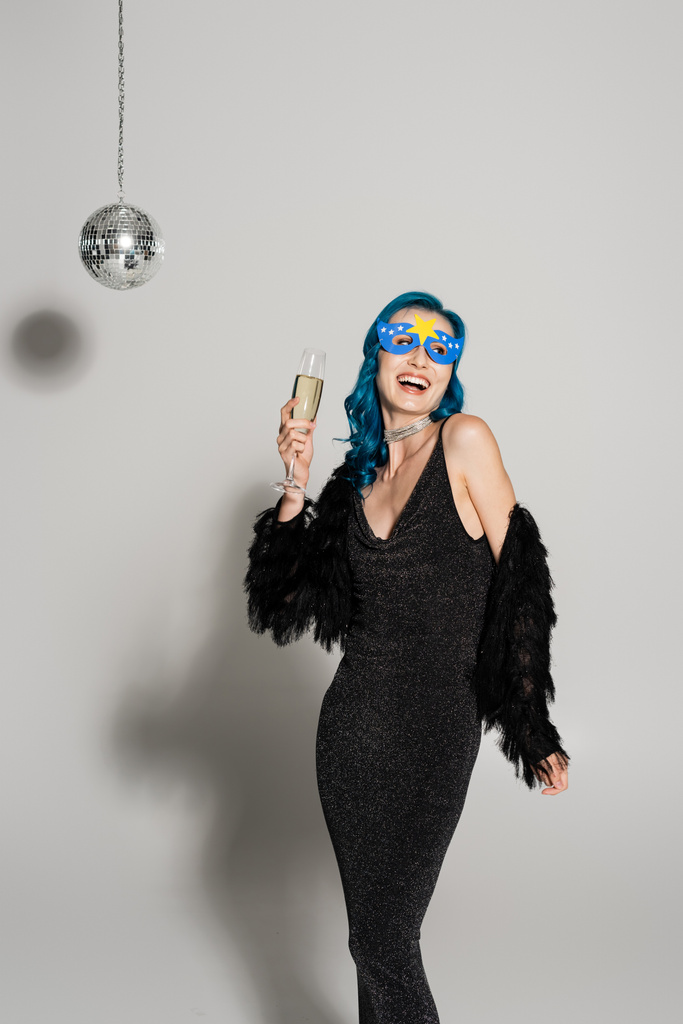 lachende vrouw in blauwe pruik en feestmasker houden champagne en weg te kijken op grijze achtergrond - Foto, afbeelding