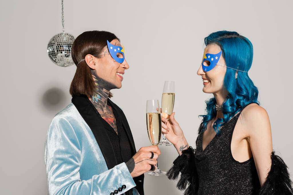 zijaanzicht van elegante niet-binaire vrienden in party maskers klinkende champagne bril op grijze achtergrond - Foto, afbeelding