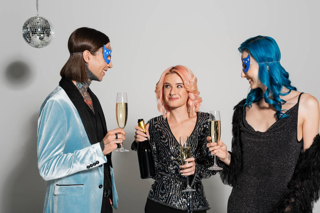 queere Person mit Champagnerflasche neben nichtbinären Freunden in Parteimasken auf grauem Hintergrund - Foto, Bild