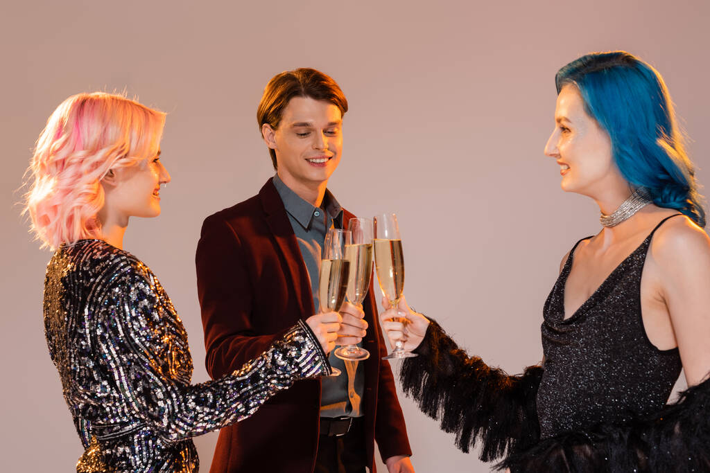 sonrientes y elegantes amigos queer tintinear copas de champán durante la fiesta de Navidad sobre fondo gris - Foto, imagen