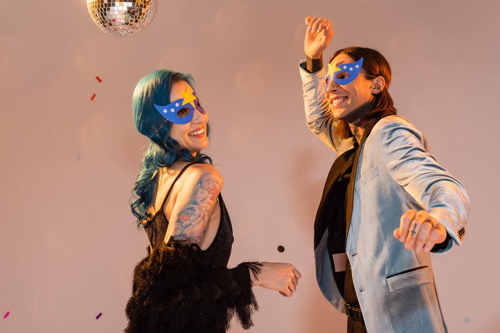 excited queer friends in stylish clothes and party masks dancing on beige background - Photo, Image
