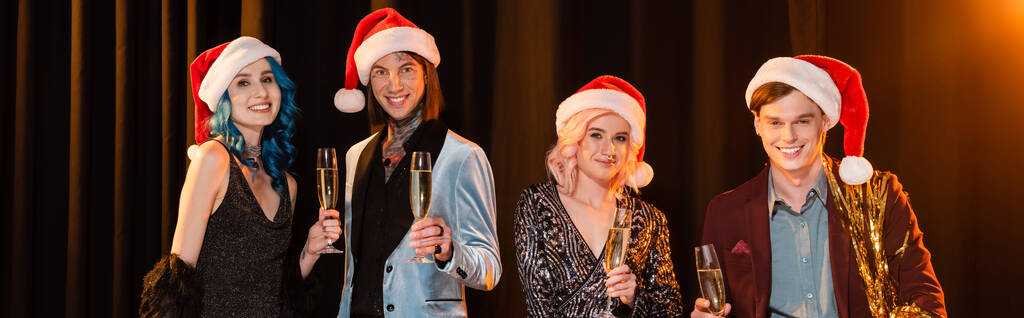 elegante nichtbinäre Freunde in Weihnachtsmannmützen lächeln in die Kamera, während sie Champagner auf dunklem Hintergrund mit Beleuchtung halten, Banner - Foto, Bild