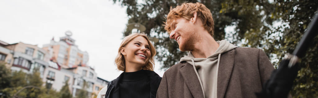 boldog vörös hajú férfi és vidám szőke nő nézik egymást, miközben sétálnak a parkban, banner - Fotó, kép
