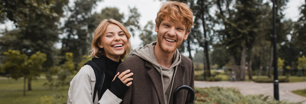 幸せなブロンド女性抱擁陽気な赤毛ボーイフレンド笑顔で秋の公園,バナー - 写真・画像