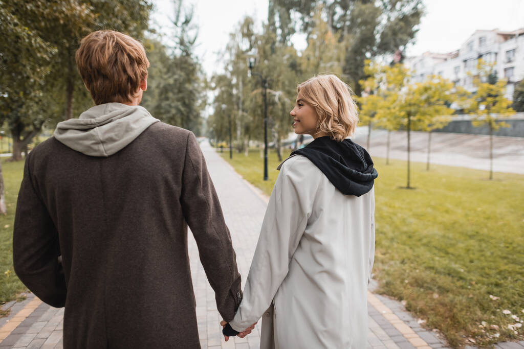 fröhliche blonde Frau schaut fröhlichen Mann beim Händchenhalten und Spazierengehen im Park an - Foto, Bild