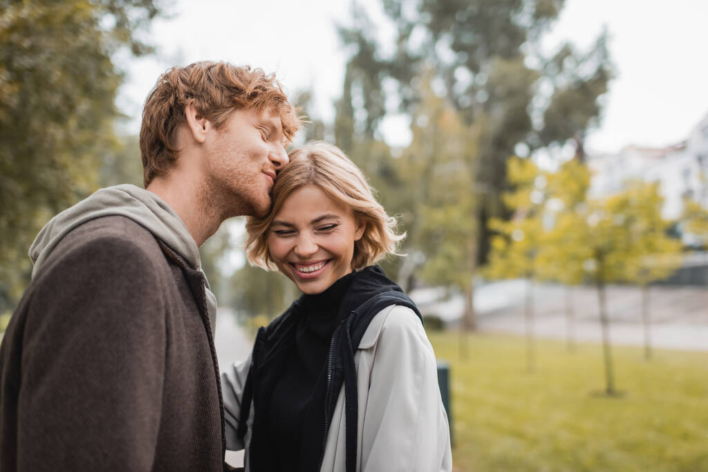 happy redhead man kissing head of blonde girlfriend smiling in park  - 写真・画像