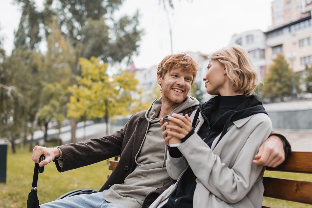 šťastný mladý muž v kabátu při pohledu na blondýny přítelkyně drží papírový šálek, zatímco sedí na lavičce - Fotografie, Obrázek