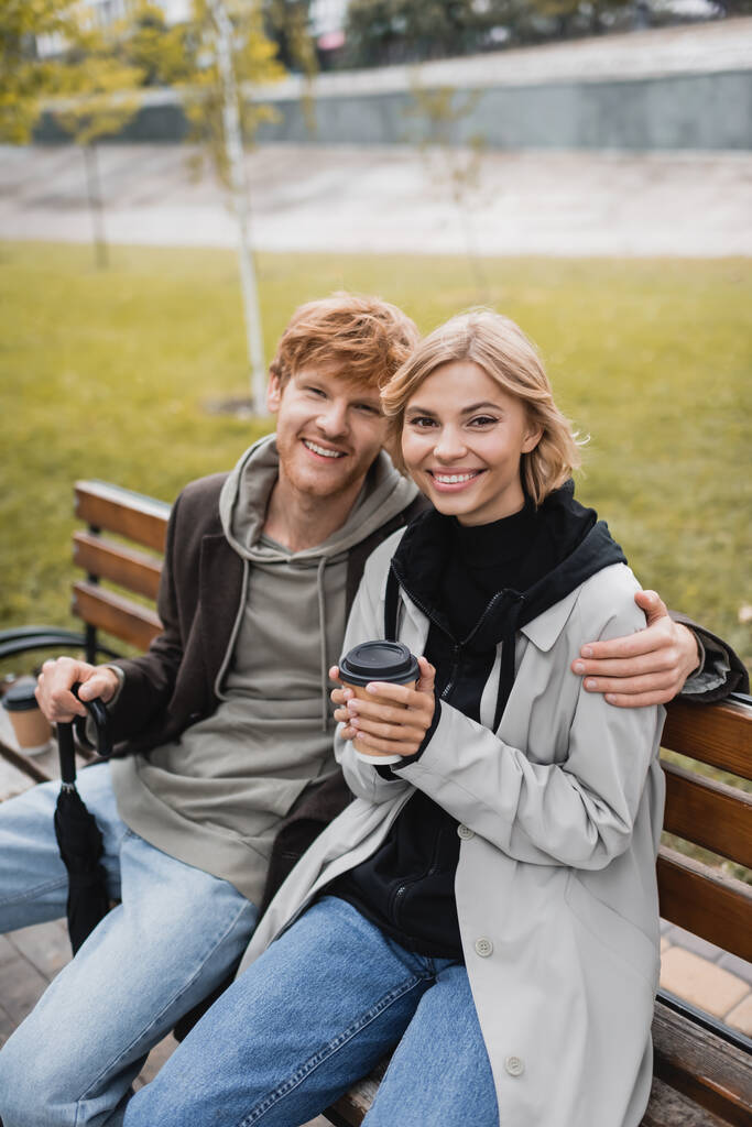 boldog fiatal férfi kabátban ölelés szőke barátnő kezében papír pohár, miközben ül a padon - Fotó, kép