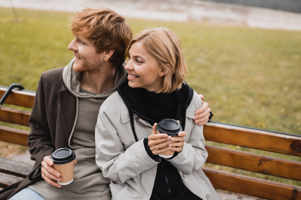 gelukkig jong paar knuffelen en vasthouden van papieren bekers met koffie te gaan terwijl zitten op de bank in park - Foto, afbeelding