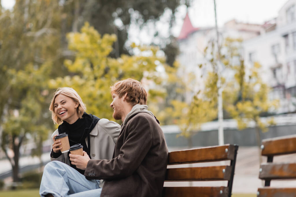 gioiosa giovane coppia ridendo e tenendo tazze di carta con caffè per andare mentre seduto sulla panchina nel parco  - Foto, immagini