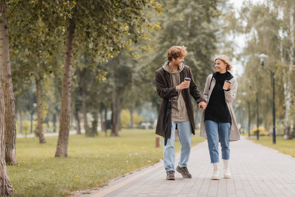 полная длина счастливой пары в пальто, держась за руки и гуляя с кофе, чтобы пойти в осенний парк  - Фото, изображение