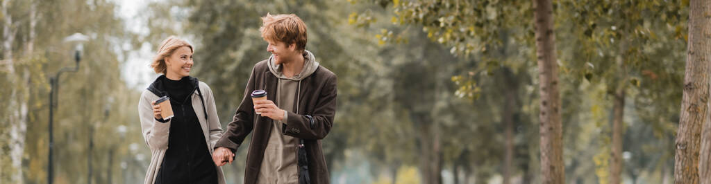 happy couple in coats holding hands and walking with paper cups in autumnal park, banner - Photo, Image
