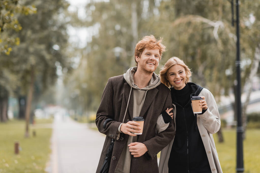 happy young couple in coats hugging and walking with paper cups in autumnal park  - Photo, Image