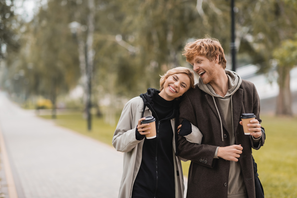 радісна молода пара в пальто обіймає і ходить з паперовими чашками в автономному парку
  - Фото, зображення