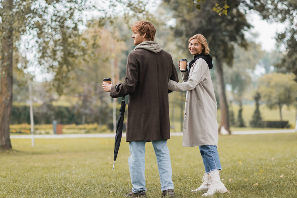 longitud completa de joven feliz en abrigo sosteniendo taza de papel y de pie con la novia en el parque otoñal  - Foto, Imagen