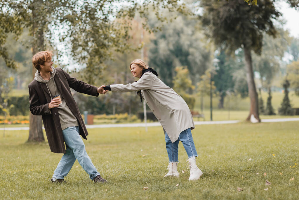pełna długość szczęśliwy młody człowiek w płaszcz gospodarstwa papierowy kubek i ciągnąc dziewczyna w jesienny park  - Zdjęcie, obraz