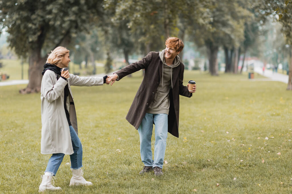 повна довжина радісного чоловіка в пальто тримає руки з усміхненою подругою в автономному парку
  - Фото, зображення