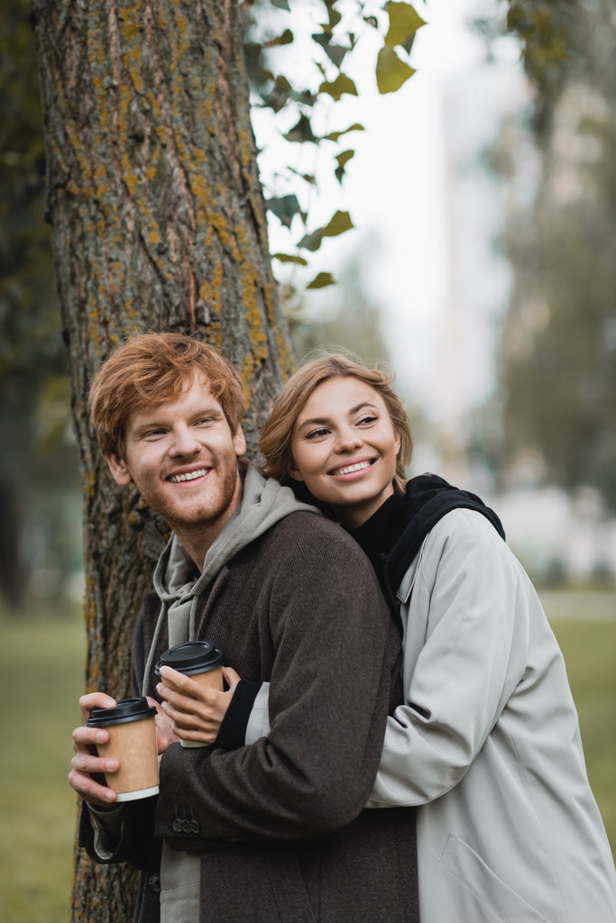 happy blonde woman holding paper cup and hugging with boyfriend near tree trunk - Photo, Image