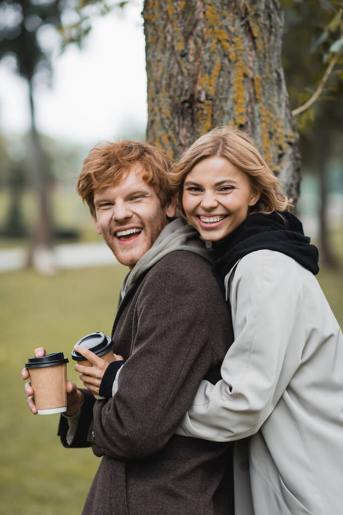 cheerful blonde woman holding paper cup and hugging with positive boyfriend near tree trunk - Photo, Image