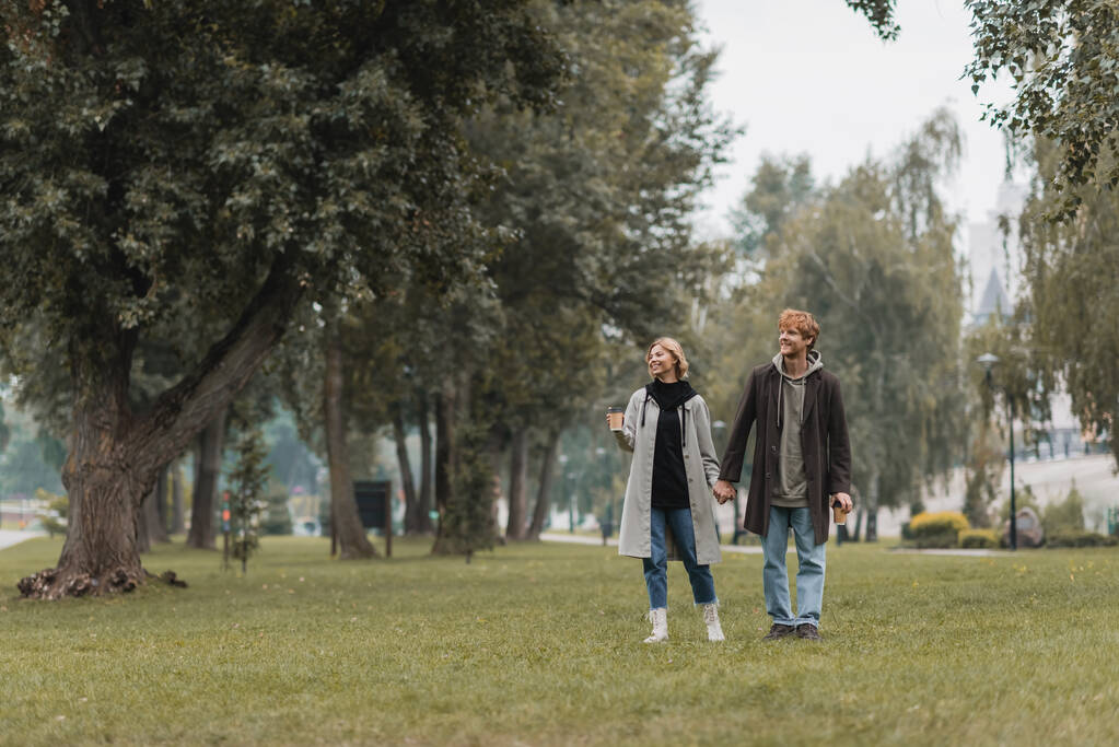 Parkta yürürken elinde kahve tutan mutlu kızıl saçlı adam ve neşeli bir kadın. - Fotoğraf, Görsel