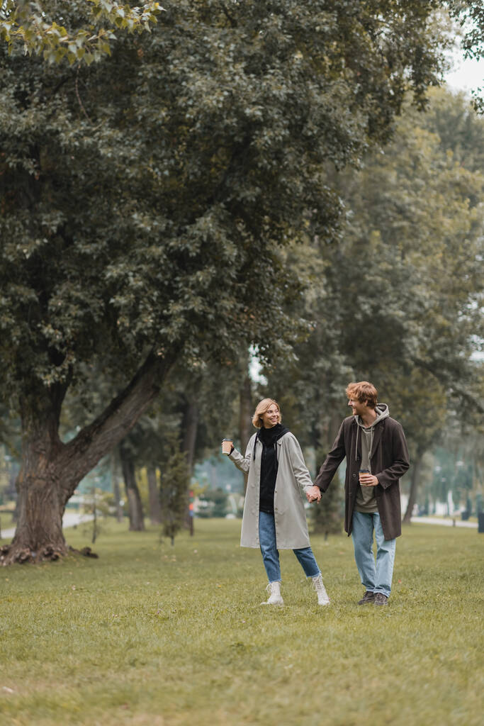 comprimento total de homem ruivo positivo e mulher alegre segurando café para ir enquanto caminha no parque - Foto, Imagem