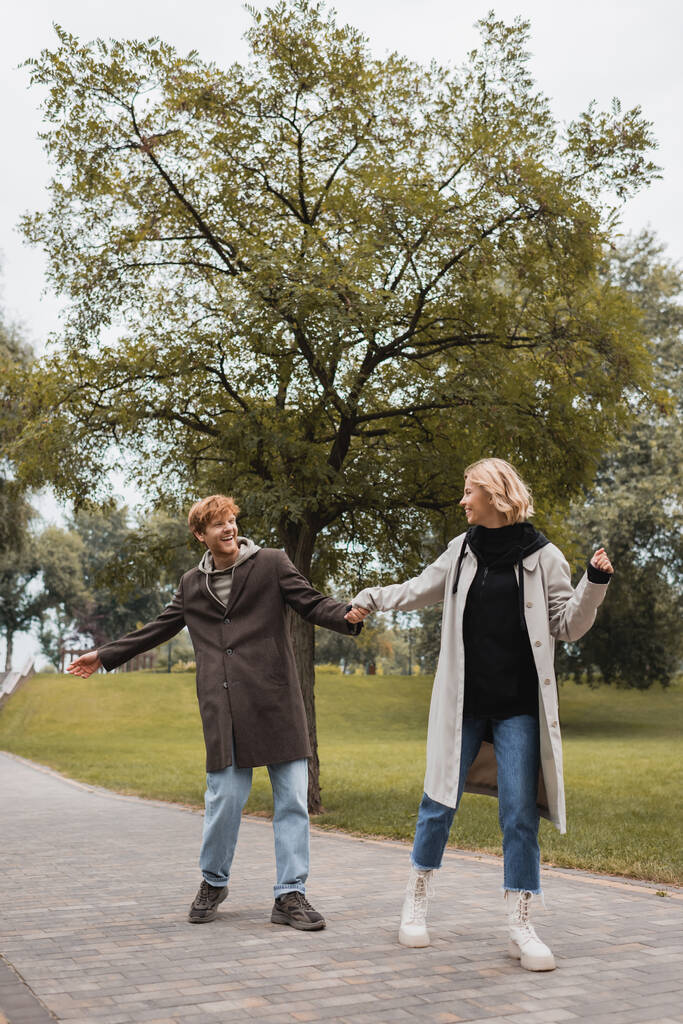 full length of happy man and pleased woman in coats holding hands while walking in park  - Photo, Image